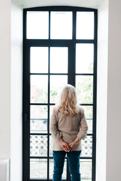 Vue arrière de la femme âgée debout près de la fenêtre à la maison pendant l'isolement — Photo de stock