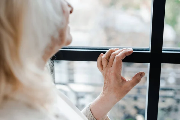 Vue recadrée de femme âgée bouleversée regardant par la fenêtre pendant l'isolement personnel — Photo de stock