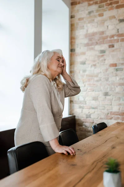 Cansado solitário sênior mulher tendo dor de cabeça e de pé à mesa durante a quarentena — Fotografia de Stock