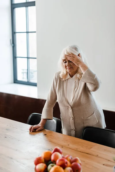 Tired elderly woman with headache standing at table with fruits during self isolation — Stock Photo