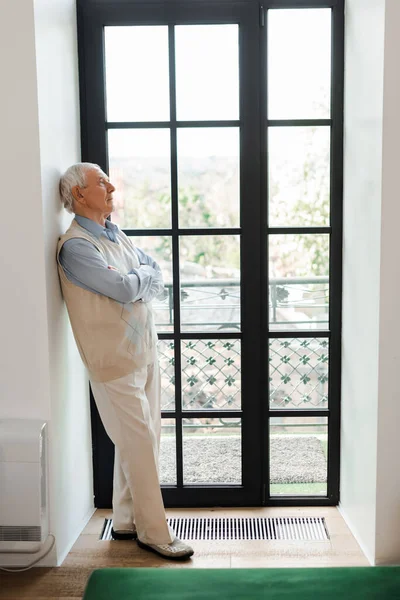 Старший человек со скрещенными руками стоит у окна во время карантина — стоковое фото