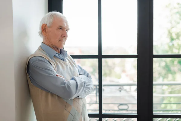 Älterer einsamer Mann mit verschränkten Armen, der während der Selbstisolierung am Fenster steht — Stockfoto