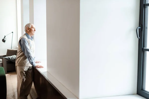 Sad elderly man standing near window during self isolation — Stock Photo