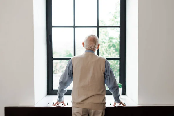 Back view of lonely elderly man standing near window during quarantine — Stock Photo