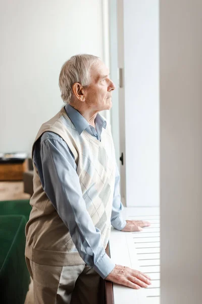 Upset elderly man standing and looking through window during quarantine — Stock Photo