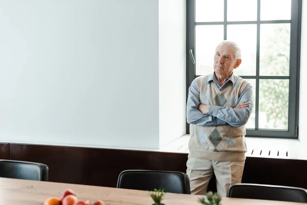 Trauriger älterer Mann steht in Selbstisolierung mit verschränkten Armen am Fenster — Stockfoto