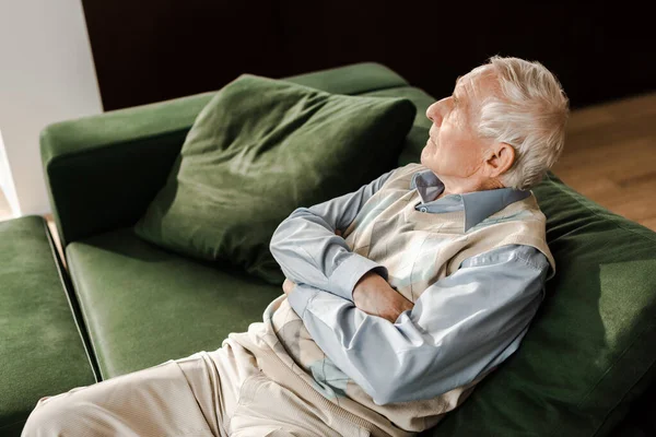 Печальный пожилой человек, сидящий со скрещенными руками на диване во время карантина — стоковое фото