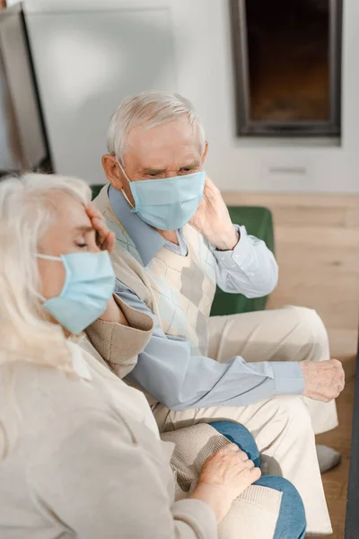 Tired senior couple in medical masks having headache while sitting on sofa during coronavirus epidemic — Stock Photo