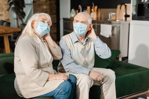 Pareja de ancianos cansados en máscaras médicas que tienen dolor de cabeza mientras están sentados en el sofá durante la epidemia de coronavirus - foto de stock