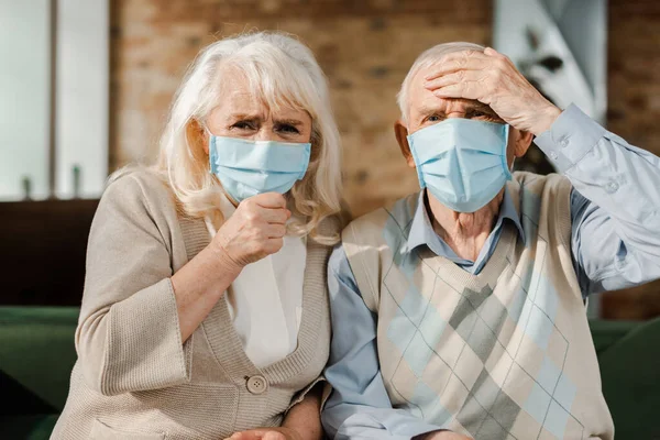 Старшая обеспокоенная пара в медицинских масках, сидящая дома во время самоизоляции — стоковое фото