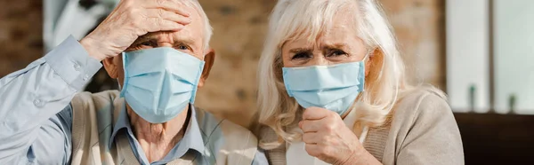 Besorgte ältere Paare in medizinischen Masken sitzen zu Hause während der Selbstisolierung, horizontales Konzept — Stockfoto