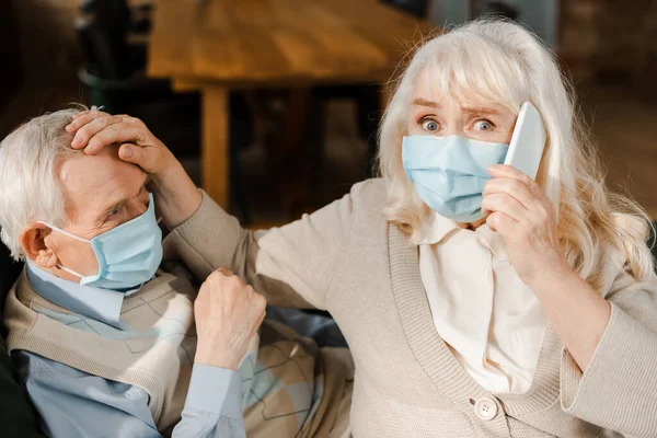 Worried elderly wife in medical mask calling doctor with smartphone while ill husband lying on sofa during coronavirus epidemic — Stock Photo