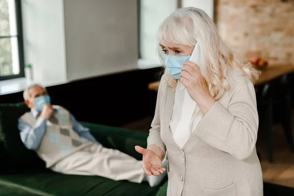 Femme âgée dans le masque médical appeler le médecin avec smartphone tandis que le mari malade couché sur le canapé pendant l'épidémie de coronavirus, foyer sélectif — Photo de stock