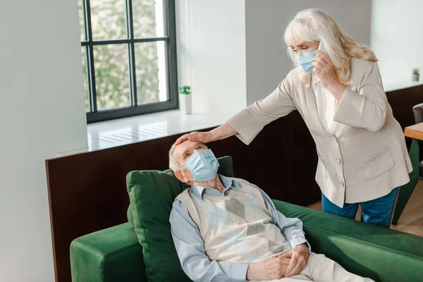 Esposa mayor en máscara médica llamando al médico con teléfono inteligente, mientras que el marido enfermo con fiebre acostado en el sofá durante la epidemia de coronavirus - foto de stock