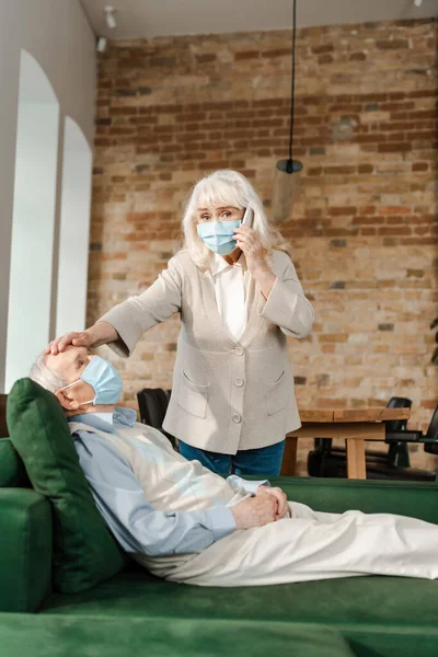 Femme âgée dans le masque médical appeler le médecin avec smartphone tandis que le mari malade avec de la fièvre couché sur le canapé pendant l'isolement personnel — Photo de stock