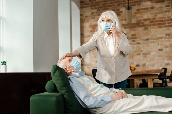 Пожилая женщина в медицинской маске вызывает врача со смартфоном, пока больной муж лежит на диване во время эпидемии коронавируса — стоковое фото