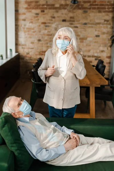 Worried elderly woman in medical mask calling doctor with smartphone while ill husband lying on sofa during quarantine — Stock Photo