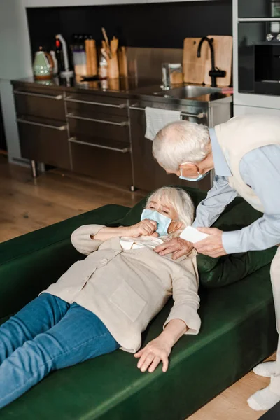 Älterer Mann in medizinischer Maske ruft Arzt mit Smartphone an, während kranke Frau während Coronavirus-Epidemie auf Sofa liegt — Stockfoto