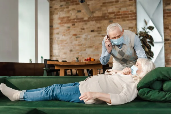 Älterer Mann mit medizinischer Maske spricht auf Smartphone, während kranke Frau während Selbstisolierung auf Sofa liegt — Stockfoto