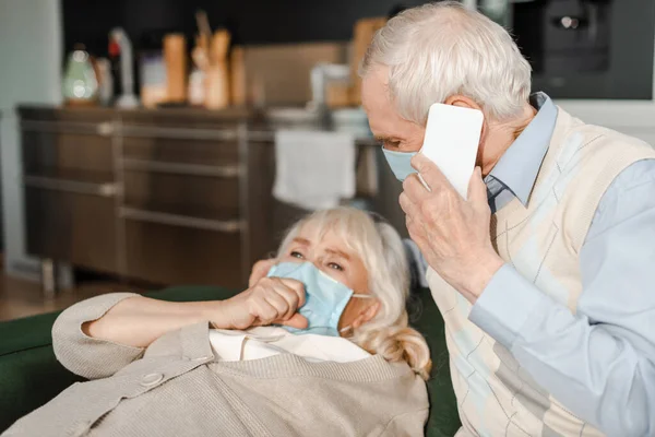 Elderly husband in medical mask calling doctor with smartphone while ill wife lying on sofa during quarantine — Stock Photo