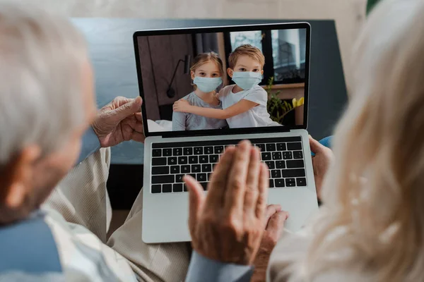 Grandparents waving while having video chat with hugging grandchildren in medical masks during self isolation — Stock Photo