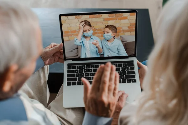 Бабушки и дедушки машут и ведут видеочат с внуками в медицинских масках, держа термометр во время карантина — стоковое фото