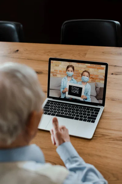 Vista ritagliata di uomo anziano mostrando pollice in su e avendo video chat con nipoti in maschere mediche con rimanere segno a casa durante l'isolamento — Foto stock