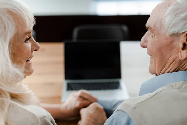 Feliz anciano pareja de ancianos tomados de la mano mientras que tiene videollamada en el ordenador portátil con pantalla en blanco en casa durante el aislamiento - foto de stock