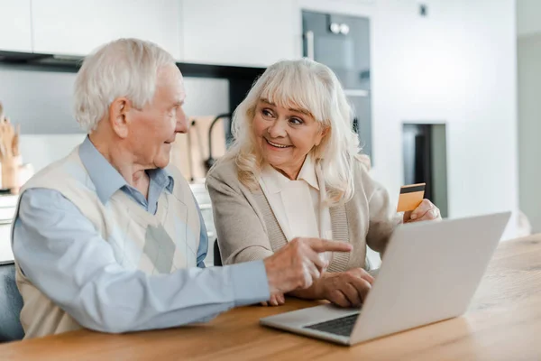 Glückliches älteres Paar kauft online mit Kreditkarte ein und zeigt während der Selbstisolierung auf den Laptop zu Hause — Stockfoto