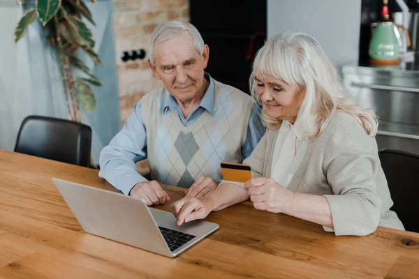 Feliz pareja de ancianos de compras en línea con tarjeta de crédito y portátil en casa durante la cuarentena - foto de stock