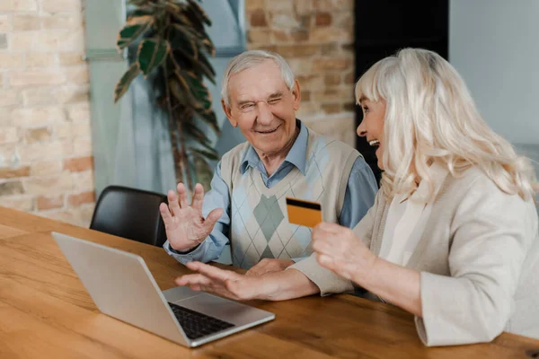 Cheerful elderly couple shopping online with credit card and laptop at home during self isolation — Stock Photo