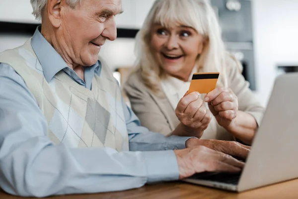 Excité couple senior shopping en ligne avec carte de crédit et ordinateur portable à la maison pendant l'isolement personnel — Photo de stock