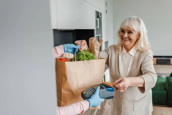 Lächelnde Seniorin, die während der Coronavirus-Pandemie mit Kreditkarte und Terminal bezahlt — Stockfoto