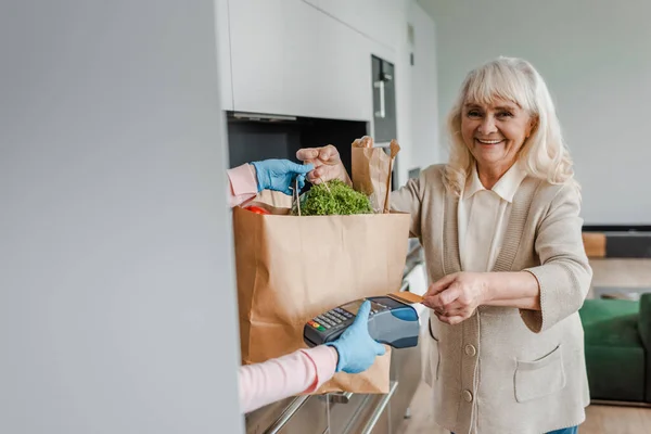 Happy elderly woman taking food delivery and paying with credit card and terminal during coronavirus pandemic — Stock Photo