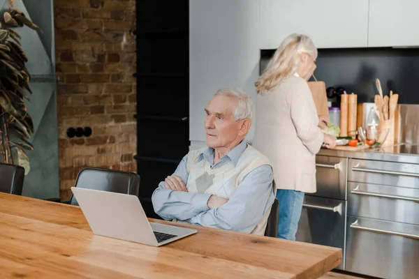Nachdenklicher Senior-Ehemann benutzt Laptop, während Ehefrau während Quarantäne in der Küche kocht — Stockfoto
