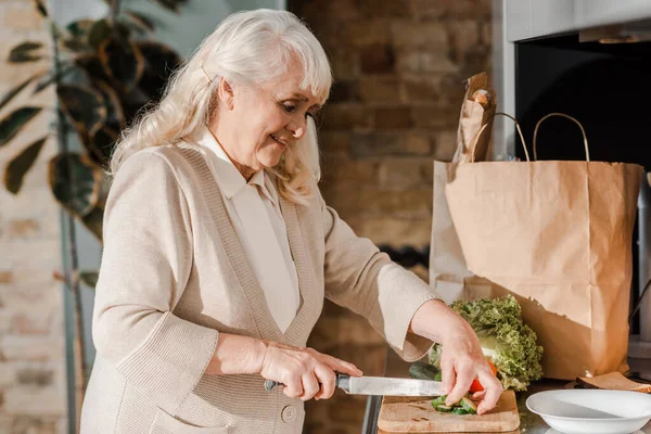 Smiling elderly woman cutting vegetables on board for salad on kitchen — Stock Photo