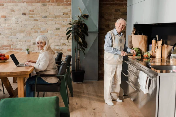 Glücklich Senior Ehemann Kochen, während Frau mit Laptop auf Küche während der Selbstisolation — Stockfoto