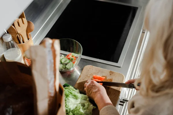 Cropped view of elderly woman cutting vegetables on board for salad on kitchen — Stock Photo