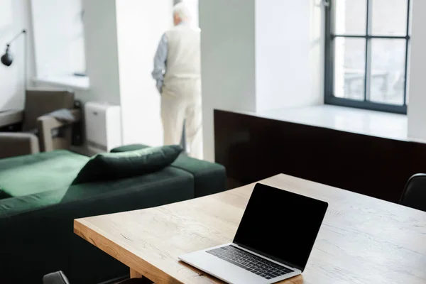 Selective focus of elderly man near window and laptop with blank screen on table — Stock Photo