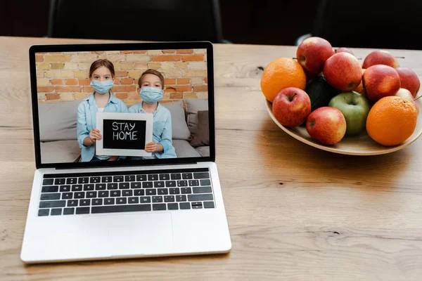 Gros plan de l'ordinateur portable avec des frères et sœurs dans des masques médicaux tenant rester signe à la maison sur l'écran sur la table avec des fruits — Photo de stock