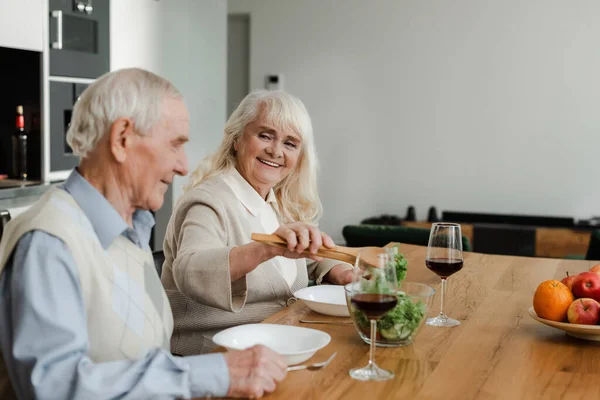 Lächelndes älteres Ehepaar beim Abendessen mit Wein und Salat zu Hause auf Selbstisolierung — Stockfoto