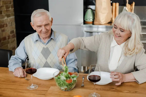 Sorrindo casal sênior jantando com vinho tinto e salada em casa em quarentena — Fotografia de Stock
