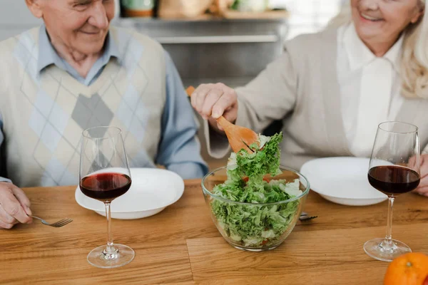 Vista recortada de feliz pareja de ancianos cenando con vino y ensalada en casa en cuarentena — Stock Photo