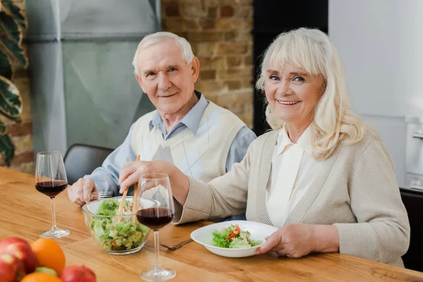 Sonriente pareja de ancianos cenando con vino y ensalada en casa en auto aislamiento - foto de stock