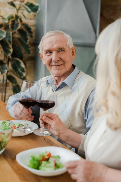 Feliz pareja de ancianos cenando con vino y ensalada en casa en cuarentena - foto de stock