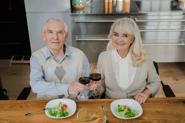 Feliz pareja de ancianos cenando con vino y ensalada en casa en auto aislamiento - foto de stock