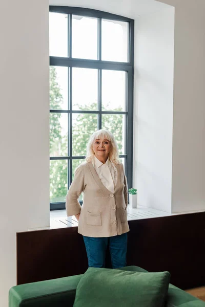 Happy elderly woman standing near window at home during self isolation — Stock Photo