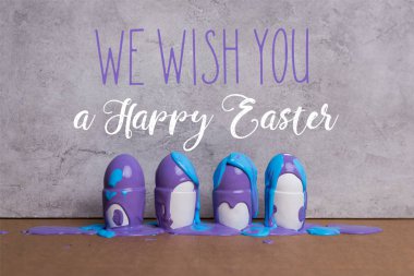 Paint covering eggs in cups with we wish you happy easter lettering on grey background clipart