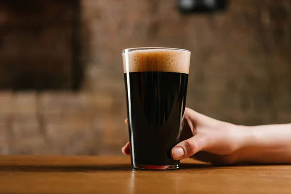 パブで新鮮な黒ビールのグラスを持っている手のショットをトリミング — ストック写真