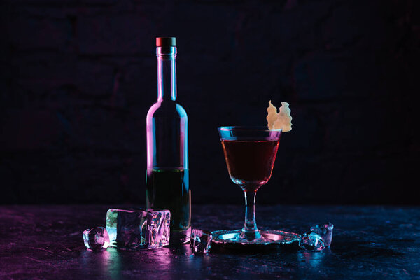 glass of alcohol cocktail, bottle of liquor and ice cubes on dark surface